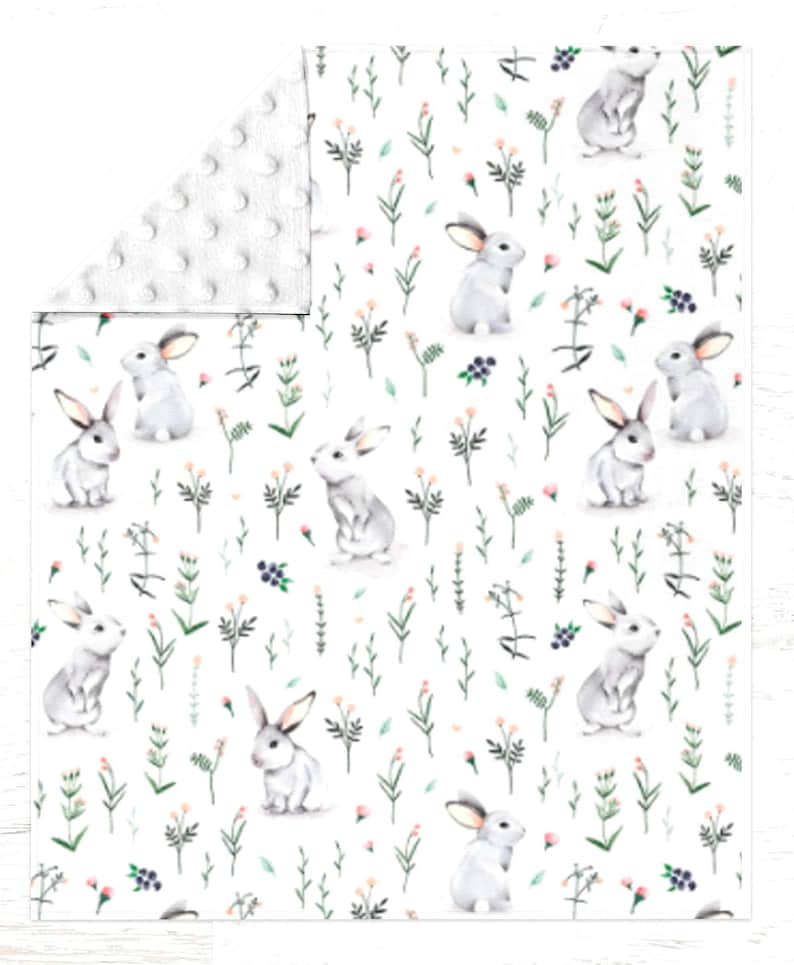 Peut inclure: Une couverture blanche avec un motif r&eacute;p&eacute;titif de lapins gris, de feuillage vert, de petites fleurs roses et de baies bleu fonc&eacute;. Le coin repli&eacute; r&eacute;v&egrave;le un tissu minky blanc textur&eacute; avec un motif de points en relief.
