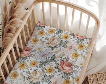 Puede incluir: Una sábana de cuna de color claro con un patrón floral repetitivo de rosas, margaritas y flores de hibisco en tonos amarillos, rosas, azules y verdes. La sábana está ajustada sobre un colchón dentro de una cuna de madera clara con barrotes.