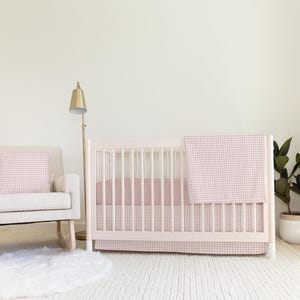 Puede incluir: Una cuna blanca con un juego de ropa de cama de cuna de vichy rosa y blanco. La cuna está en una guardería con una mecedora blanca y una planta en maceta.