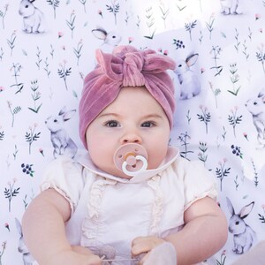 Peut inclure: Un turban pour b&eacute;b&eacute; en velours mauve avec un n&oelig;ud sur le devant, port&eacute; avec une barboteuse &agrave; volants cr&egrave;me &agrave; manches courtes, une sucette rose p&acirc;le et des chaussettes en tricot c&ocirc;tel&eacute;, sur un fond en tissu blanc &agrave; motifs de lapins et fleurs.