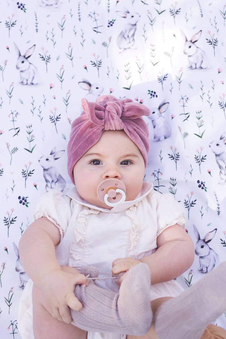 Peut inclure: Un turban pour b&eacute;b&eacute; en velours mauve avec un n&oelig;ud sur le devant, port&eacute; avec une barboteuse &agrave; volants cr&egrave;me &agrave; manches courtes, une sucette rose p&acirc;le et des chaussettes en tricot c&ocirc;tel&eacute;, sur un fond en tissu blanc &agrave; motifs de lapins et fleurs.