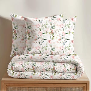 Könnte beinhalten: Ein Bettwäscheset mit weißem Hintergrund und einem sich wiederholenden Aquarellmuster aus zartrosa Rosen, grünen Blättern und kleinen Kolibris. Das Set enthält zwei quadratische Kissenbezüge und eine gefaltete Bettdecke mit demselben Blumenmotiv.