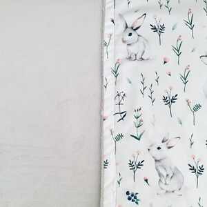 Peut inclure: Une couverture en tissu blanc avec un motif r&eacute;p&eacute;titif de lapins gris clair, de petites fleurs roses, de tiges vertes et de baies bleu fonc&eacute;. Le tissu pr&eacute;sente un aspect doux et textur&eacute; avec une bordure cousue le long de l'imprim&eacute; lapins.