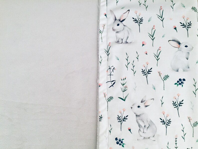 Peut inclure: Une couverture en tissu blanc avec un motif r&eacute;p&eacute;titif de lapins gris clair, de petites fleurs roses, de tiges vertes et de baies bleu fonc&eacute;. Le tissu pr&eacute;sente un aspect doux et textur&eacute; avec une bordure cousue le long de l'imprim&eacute; lapins.