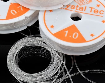 May include: Close-up of several spools of clear, elastic beading cord. The spools are white with orange labels indicating the cord's diameter in millimeters. One spool is labeled "1.0", and another is labeled "0.7". The cord is coiled and ready for use.