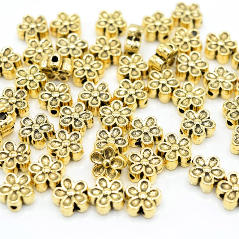 Peut inclure: Un grand groupe de perles en m&eacute;tal de couleur dor&eacute;e en forme de fleur. Les perles sont &eacute;parpill&eacute;es sur une surface blanche.