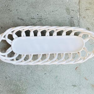 May include: A white, oval-shaped decorative basket with a woven design. The basket has a solid, rectangular center and a braided border. This item is suitable for home decor and storage.