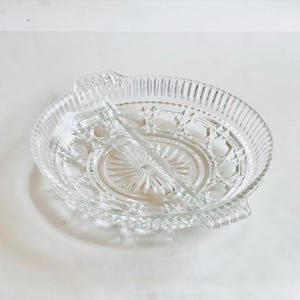 May include: Clear glass serving dish with two compartments. The dish has a circular shape with a decorative starburst pattern in the center and a ribbed edge. It features two handles and a divider.