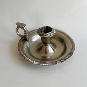 May include: A silver-colored candlestick holder with a round base and a handle. The candle holder has a circular base with a raised, textured rim. The candle cup is in the center, and the handle is on the side.