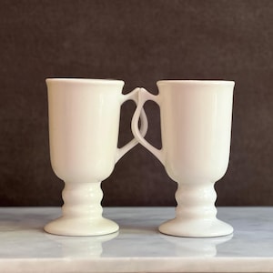 May include: Two white ceramic mugs with a unique handle design, joined in the middle. The mugs have a pedestal base and a smooth, glossy finish. They are set against a neutral background, highlighting their elegant form.