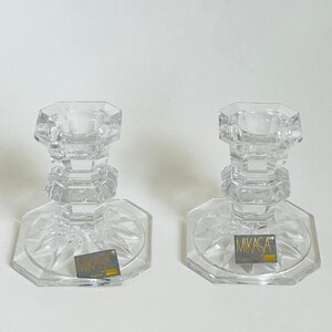 May include: Two clear glass candle holders with a geometric design. Each holder has a round base with a raised center and a square-shaped top. The base of each holder has a small label that reads "Mikasa Crystal".