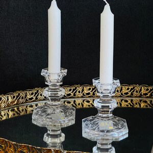 May include: Two clear glass candle holders with white candles on a gold tray. The candle holders have a hexagonal base and a round top. The tray has a decorative gold border.