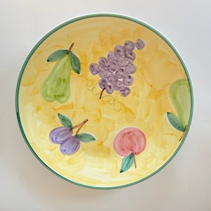 May include: A round, hand-painted ceramic plate with a yellow background and a green rim. The plate features illustrations of green pears, purple grapes, plums, and a red peach. The fruit illustrations are in a whimsical style.
