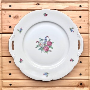 May include: A white porcelain serving platter with a floral design featuring pink and white roses and blue forget-me-nots. The platter has two handles and a gold rim.