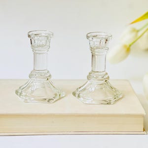 May include: Two clear glass candle holders with a classic design. The candle holders are placed on a closed book with a light-colored cover. White tulips are in the background. The candle holders are suitable for home decor.