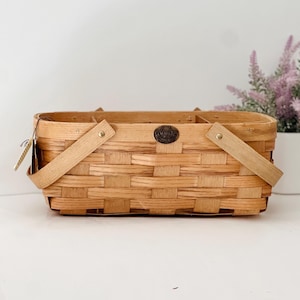 May include: A rectangular, light brown woven basket with wooden handles and a metal emblem. The basket has a tag attached to the side. The basket is made of woven wood strips.
