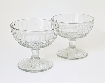May include: Two clear glass dessert bowls with a pedestal base. The bowls have a textured, fluted design.