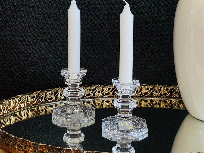 May include: Two clear glass candle holders with white candles on a gold tray. The candle holders have a hexagonal base and a round top. The tray has a decorative gold border.