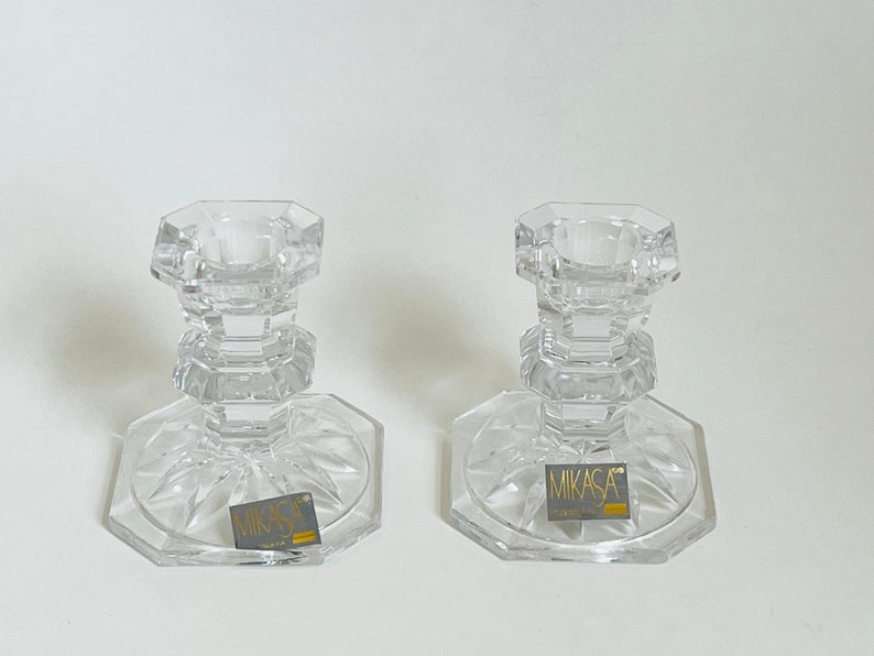 May include: Two clear glass candle holders with a geometric design. Each holder has a round base with a raised center and a square-shaped top. The base of each holder has a small label that reads "Mikasa Crystal".