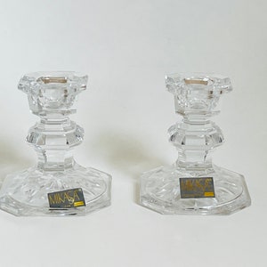 May include: Two clear glass candle holders with a cut glass design. The holders have a square base and a round top. The base of each holder has a gold label that reads "Mikasa".