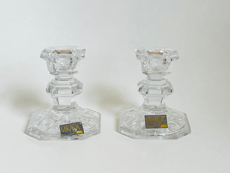 May include: Two clear glass candle holders with a cut glass design. The holders have a square base and a round top. The base of each holder has a gold label that reads "Mikasa".