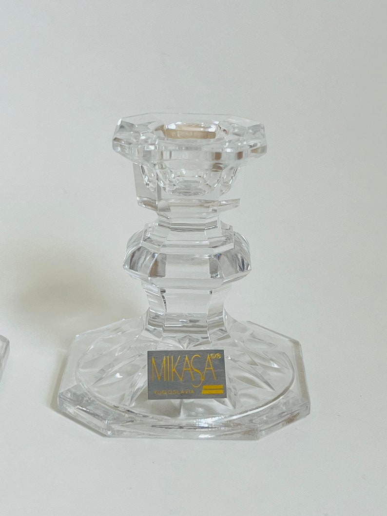 May include: A clear glass candle holder with a cut crystal design. The base of the holder has a gold label that reads "Mikasa Crystal of Japan".