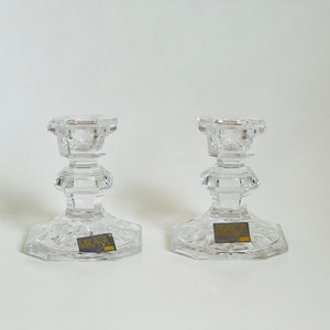 May include: Two clear glass candle holders with a cut glass design. The holders have a square base and a round top. The base of each holder has a gold label that reads "Mikasa".