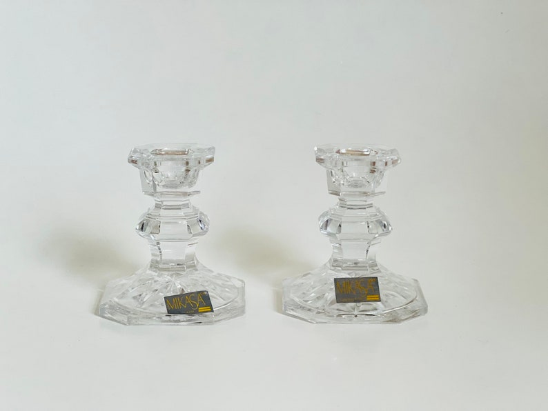 May include: Two clear glass candle holders with a cut glass design. The holders have a square base and a round top. The base of each holder has a gold label that reads "Mikasa".