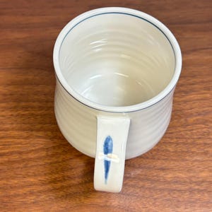 Puede incluir: Taza de cer&aacute;mica color crema con borde azul y dise&ntilde;o de pez azul en el asa. La taza tiene una textura acanalada y un dise&ntilde;o simple y elegante. Un complemento perfecto para cualquier cocina o colecci&oacute;n de caf&eacute;.