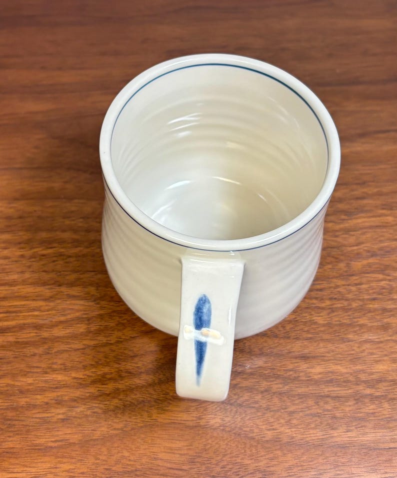 Puede incluir: Taza de cer&aacute;mica color crema con borde azul y dise&ntilde;o de pez azul en el asa. La taza tiene una textura acanalada y un dise&ntilde;o simple y elegante. Un complemento perfecto para cualquier cocina o colecci&oacute;n de caf&eacute;.