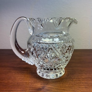 May include: A clear, cut-glass pitcher with a curved handle and a decorative rim. The pitcher features a diamond pattern on the body and a starburst design on the base. The pitcher is sitting on a wooden surface.