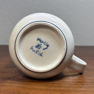 Puede incluir: Taza de cer&aacute;mica blanca con cuerpo redondeado y asa peque&ntilde;a. La base de la taza presenta el texto manuscrito "Molly Hatch" en tinta azul, junto con un peque&ntilde;o dise&ntilde;o de corona. Una fina l&iacute;nea azul rodea el borde.