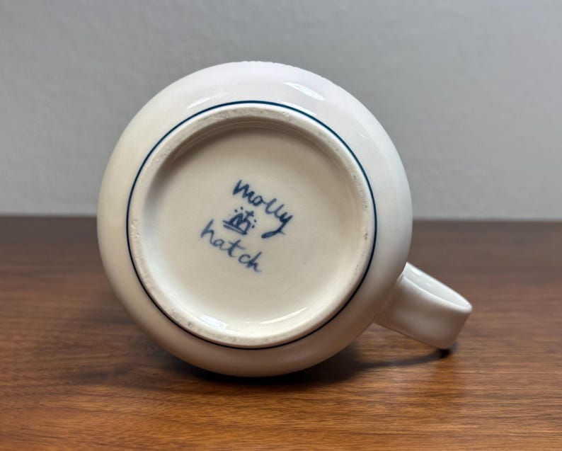 Puede incluir: Taza de cer&aacute;mica blanca con cuerpo redondeado y asa peque&ntilde;a. La base de la taza presenta el texto manuscrito "Molly Hatch" en tinta azul, junto con un peque&ntilde;o dise&ntilde;o de corona. Una fina l&iacute;nea azul rodea el borde.