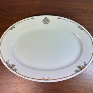 Könnte beinhalten: Ovale weiße Servierplatte mit dekorativem Rand. Der Rand hat eine dünne braune Linie mit kleinen floralen und Eichel-Akzenten. Oben ist ein Wappen zentriert. Ein klassisches Stück zum Servieren von Speisen.