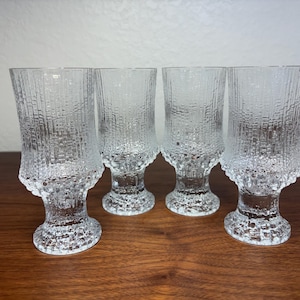 以下が含まれることがあります： ユニークな垂直線模様の、透明で質感のあるガラスゴブレット4つ。ゴブレットは台座とわずかにフレアしたリムを備えています。グラスは濃い木製の表面に配置され、そのデザインと透明度を際立たせています。