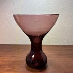 Può includere: Un vaso di vetro color ametista vintage. Il vaso ha una parte superiore ampia e svasata che si assottiglia fino a un collo stretto e una base arrotondata. Il vetro ha una finitura liscia e lucida e il vaso è posto su una superficie di legno.