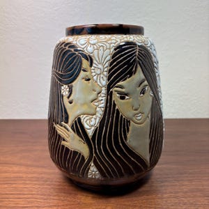 May include: A brown ceramic vase with a detailed relief design. The vase features two stylized female figures with long hair, set against a backdrop of white floral patterns. The rim and base are a darker brown.