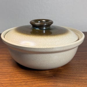 May include: A ceramic bowl with a lid. The bowl is a light beige colour with a speckled texture. The lid is a darker brown colour with a rounded handle. The bowl is on a wooden surface.