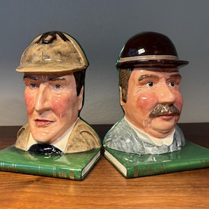 May include: A white ceramic mug with a black handle and the text "But First, Coffee" in a bold, black font. The mug is set against a blurred background, suggesting a cozy setting. The mug appears to be approximately 4 inches tall.