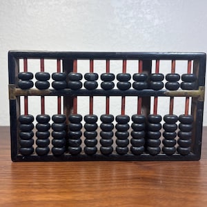 May include: A vintage abacus with a black wooden frame and black beads. The beads are arranged on red rods. The abacus has brass-colored metal accents and sits on a wooden surface.