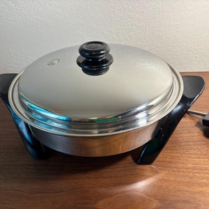 Puede incluir: Una sartén eléctrica de acero inoxidable con tapa y asas negras. La tapa tiene un pomo negro. La sartén está sobre una superficie de madera. Se ve un cable de alimentación negro y un panel de control.