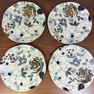 May include: Four white ceramic plates with a floral design. Each plate features a large, central rose in shades of gold and brown, surrounded by gray and black leaves and smaller white flowers. The plates are arranged on a wooden surface.