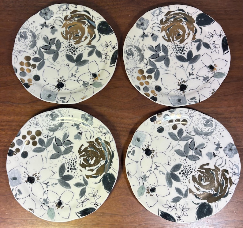 May include: Four white ceramic plates with a floral design. Each plate features a large, central rose in shades of gold and brown, surrounded by gray and black leaves and smaller white flowers. The plates are arranged on a wooden surface.