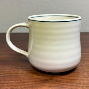 Puede incluir: Una taza de cer&aacute;mica color crema con base redondeada y asa curva. La taza tiene una l&iacute;nea azul oscuro alrededor del borde. El cuerpo de la taza tiene estr&iacute;as horizontales. La taza est&aacute; sobre una superficie de madera.