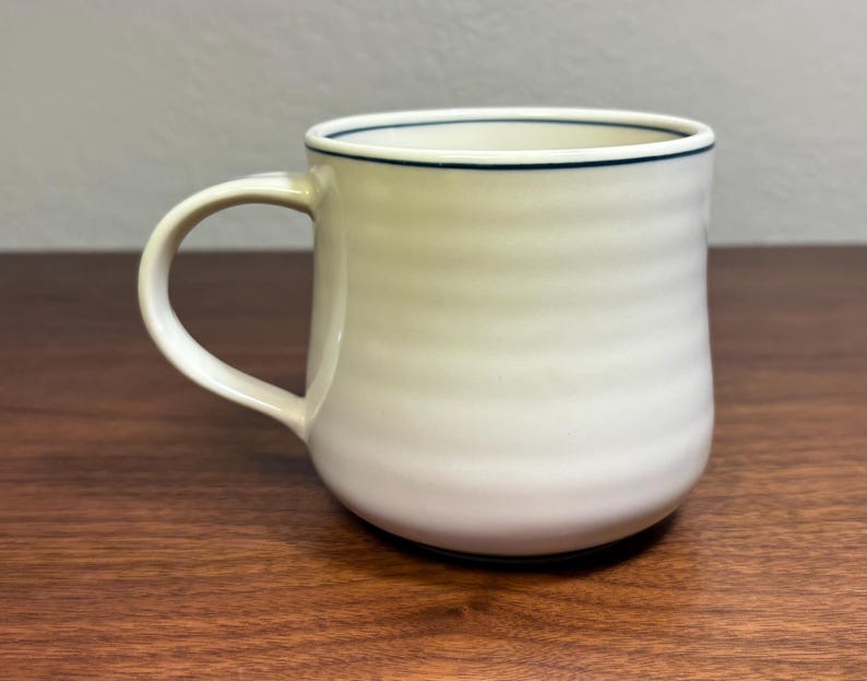 Puede incluir: Una taza de cer&aacute;mica color crema con base redondeada y asa curva. La taza tiene una l&iacute;nea azul oscuro alrededor del borde. El cuerpo de la taza tiene estr&iacute;as horizontales. La taza est&aacute; sobre una superficie de madera.