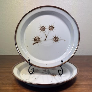 Pode incluir: Dois pratos de cerâmica branca com borda marrom e design de estrela marrom. Os pratos são exibidos em um suporte de metal preto, mostrando sua estética vintage. Os pratos são de cor neutra, tornando-os versáteis para qualquer mesa.