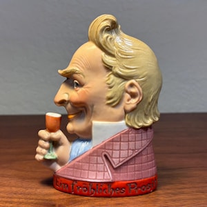 May include: A ceramic figurine in the shape of a man's head, holding a small glass. The man has blonde hair and is wearing a pink jacket with a diamond pattern. The base of the figurine has the words "Ein fröhliches Prost" in red.