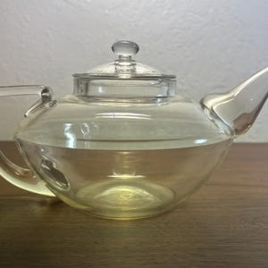 Puede incluir: Una tetera de vidrio transparente con asa y boquilla curvadas. La tetera tiene una tapa con un pequeño pomo y está llena de un líquido de color claro. La tetera está sobre una superficie de madera.