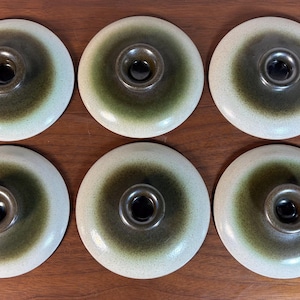 May include: Six ceramic lids with a central, dark-coloured knob. The lids have a circular shape with a light-coloured outer ring and a darker, olive-green centre. The knobs are a darker shade of brown. The lids are arranged on a wooden surface.