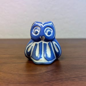 Może przedstawiać: Mała ceramiczna figurka sowy w odcieniach niebieskiego i białego. Sowa ma duże, okrągłe oczy i stylizowany wzór piór na piersi. Figurka stoi na drewnianej powierzchni.